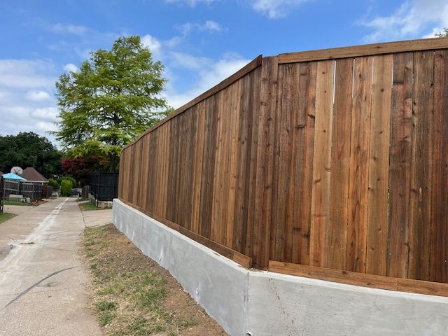 Brown wooden fence on a concrete base, bordering a sidewalk and yard, under a partly cloudy sky.