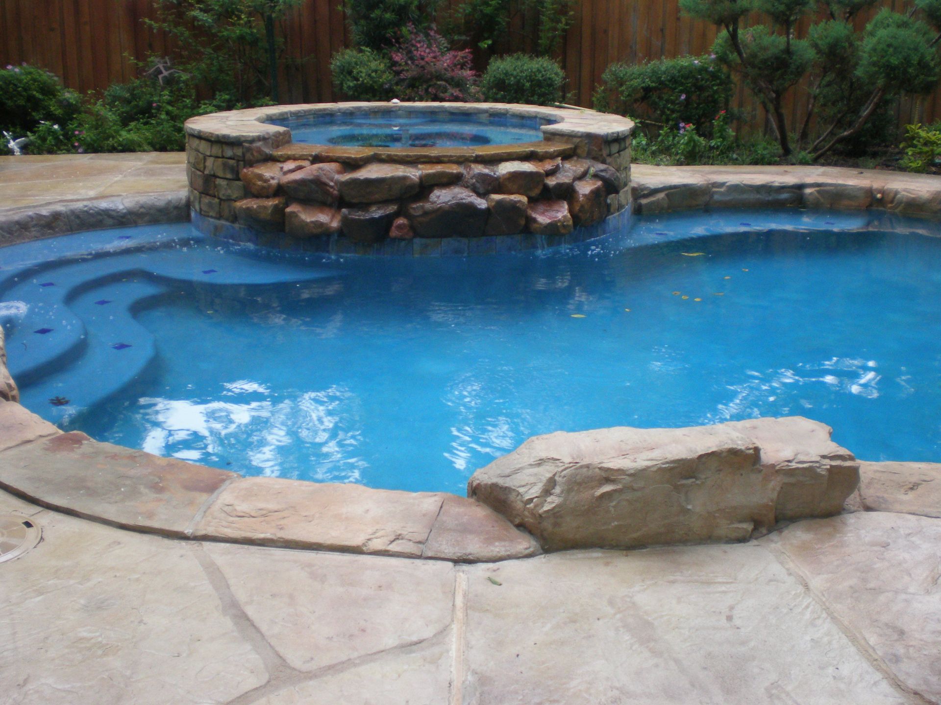 Swimming pool with built-in hot tub and stone waterfall feature, surrounded by stone patio and lush greenery.