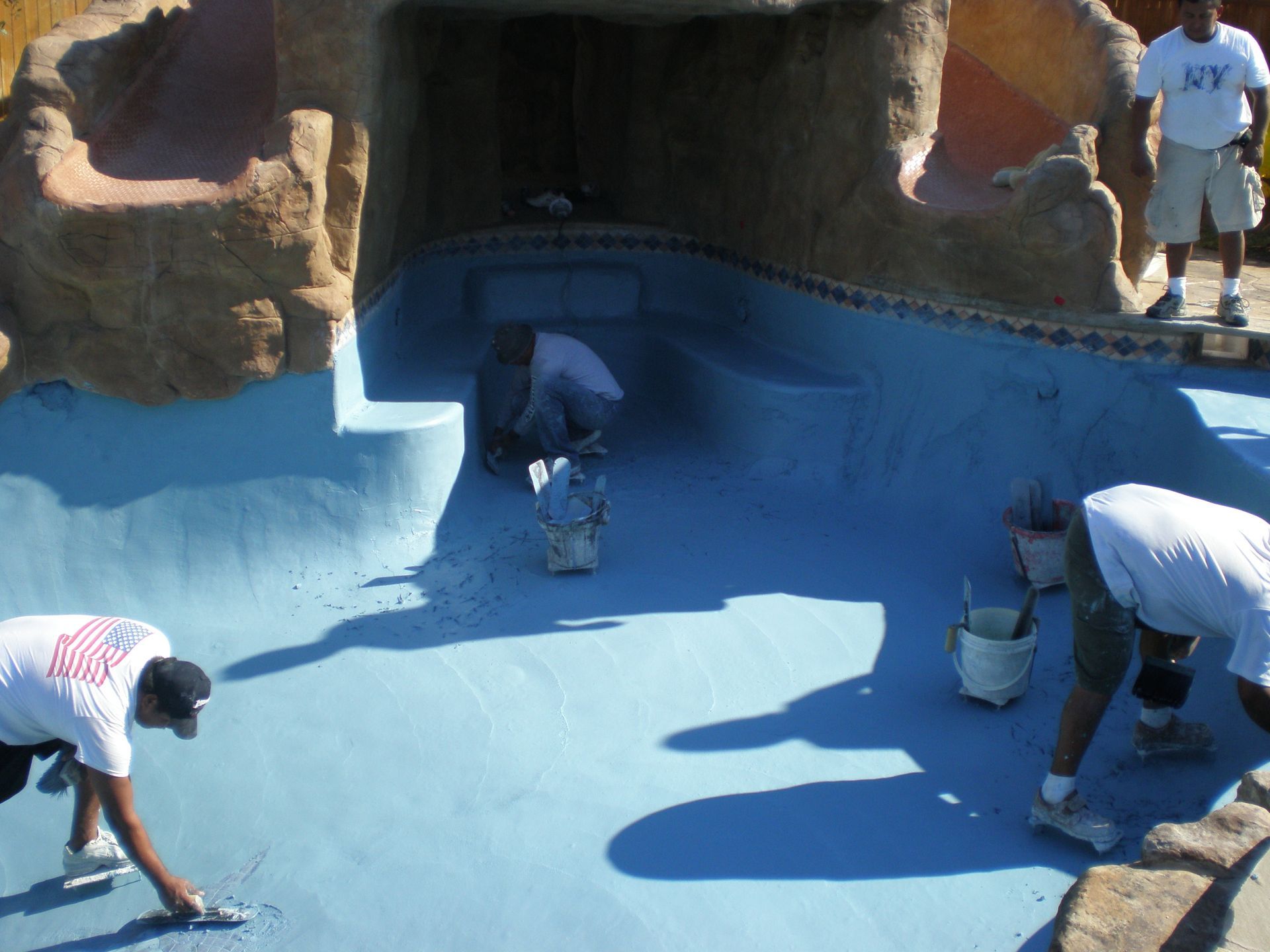 Men in white shirts work on the interior of a blue swimming pool, applying what looks like plaster.