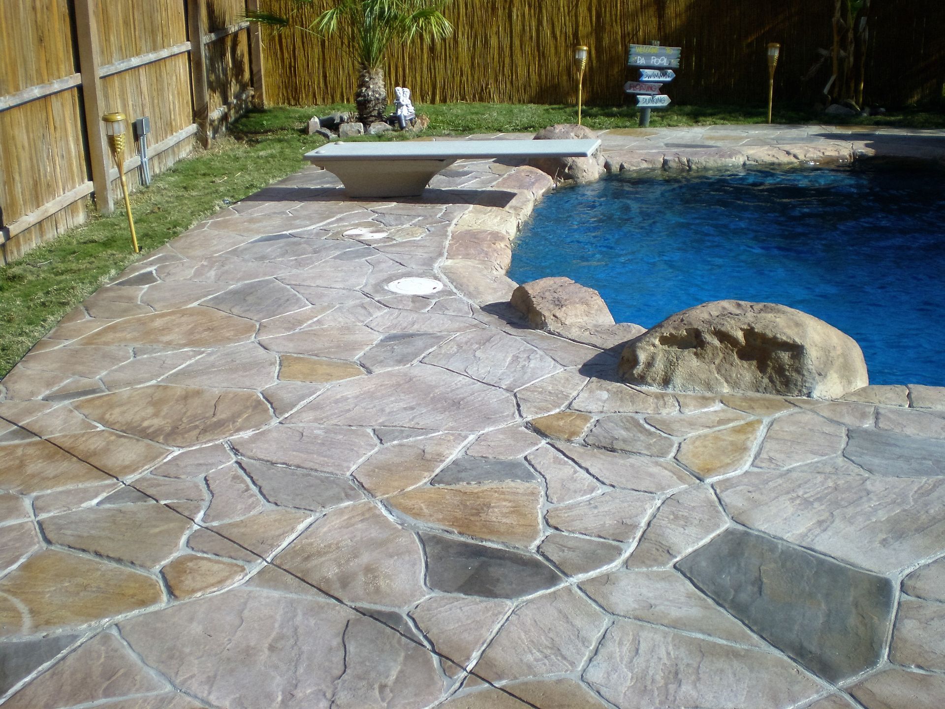 Concrete pool deck with stone pattern, blue water, diving board, and surrounding fence.
