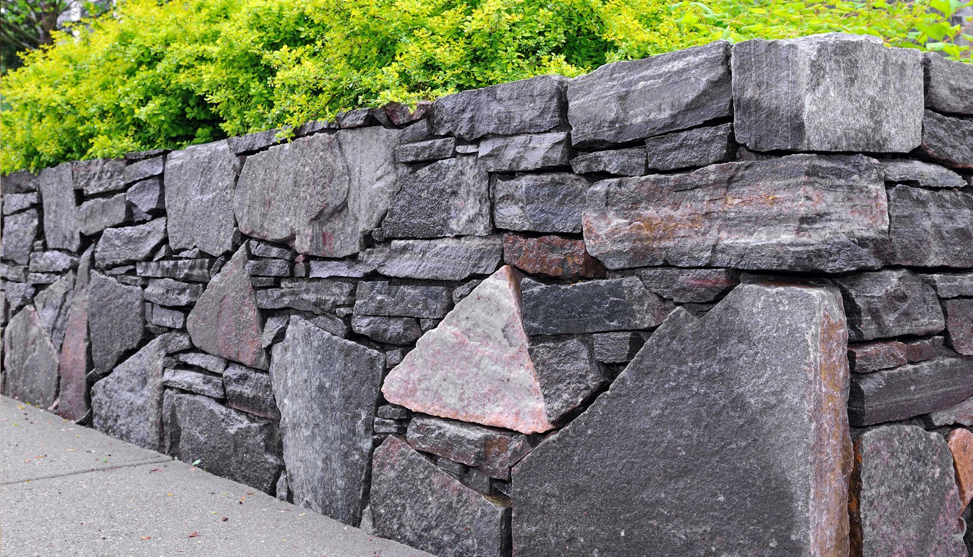 Stone wall with varying sizes and shapes, gray and brown tones, with green foliage in the background.