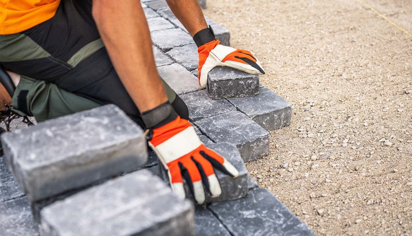 A construction worker wearing gloves laying gray paving stones on a sandy surface.