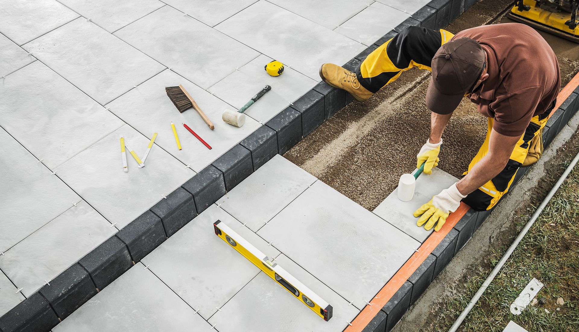 Man laying concrete patio tiles, using tools on a gravel base, with bordering bricks and landscaping.