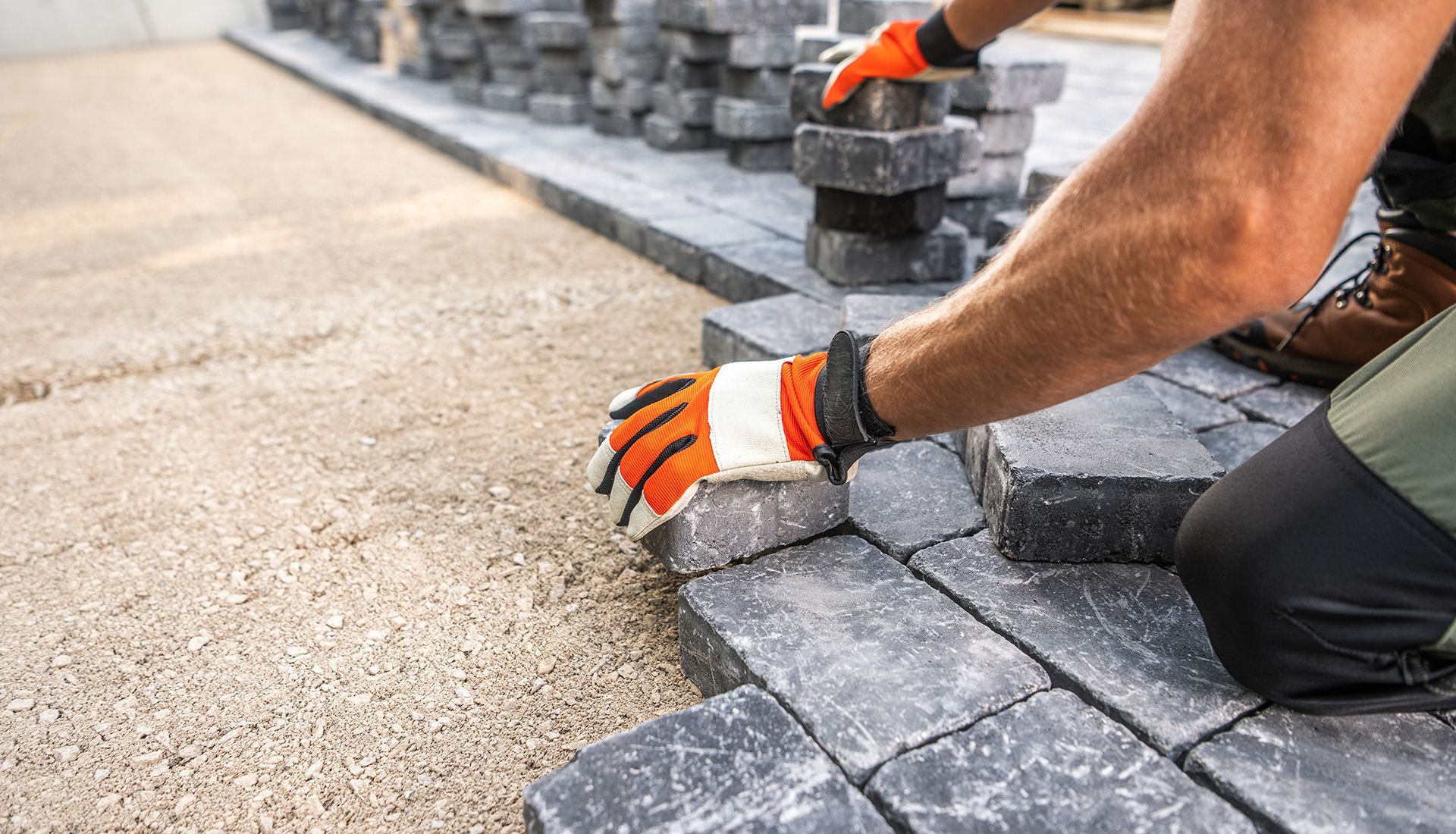 Construction worker laying dark gray paving stones, wearing orange gloves.