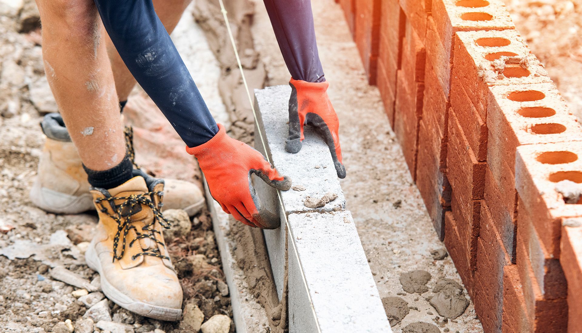 Person laying a concrete block, wearing gloves and boots, building a wall outdoors.