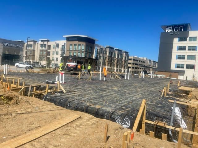 Construction site with groundwork covered in black sheeting; workers nearby, buildings in background, bright blue sky.
