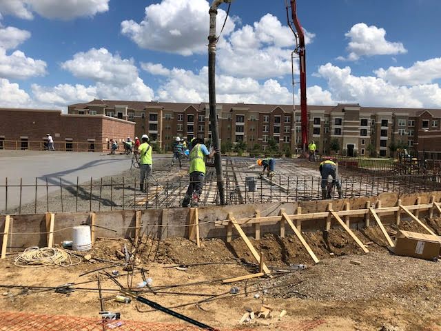 Construction site with workers pouring concrete. Rebar framework, wooden forms, and a concrete pump truck are visible.