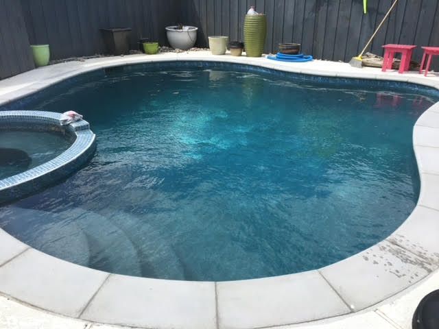 A small backyard swimming pool with a jacuzzi, surrounded by paving and a dark wooden fence.
