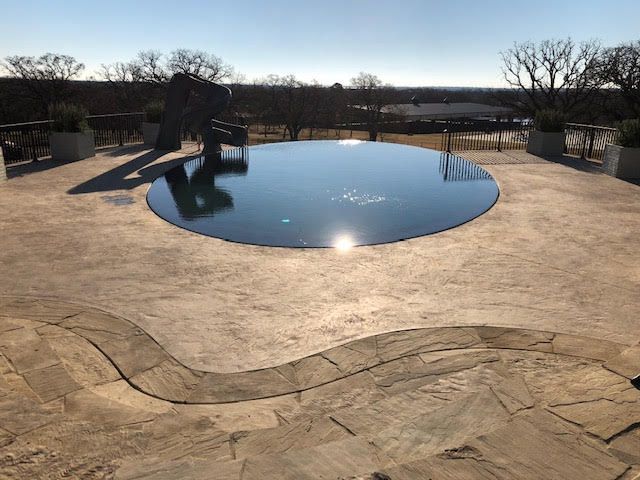 Circular pool on a patio, reflecting the sky, with a sculpture and trees in the background.