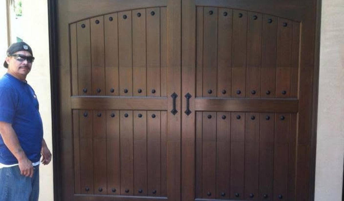 Person standing beside large dark wooden double doors with metal handles and hinges.