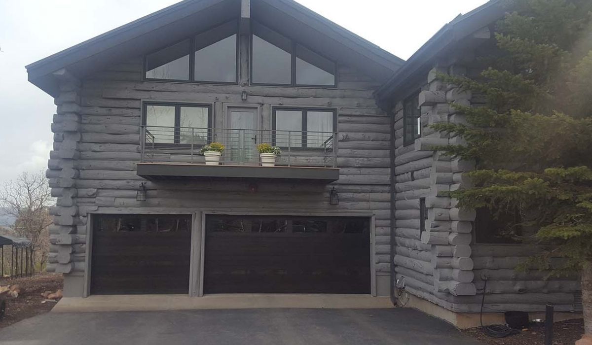 Gray log-style house with two garage doors, balcony, and stone accents under an overcast sky