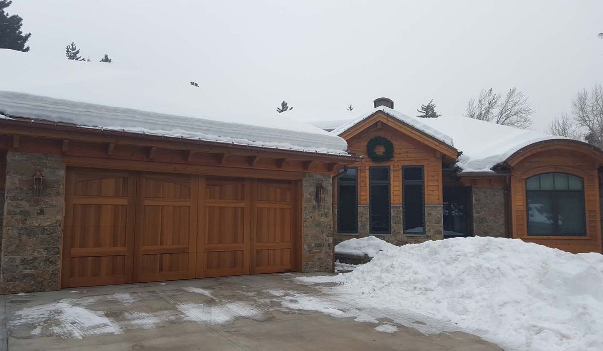 Snowy house exterior with wooden garage doors and stone siding under an overcast sky