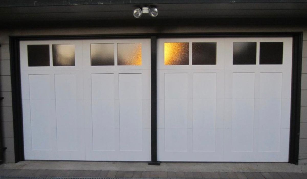 Two white garage doors with small windows, lit warmly from inside at dusk