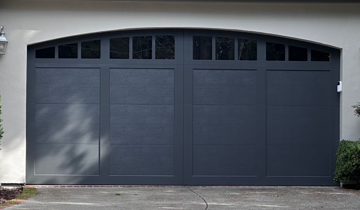 Dark gray double garage door with arched window panels on a white house exterior