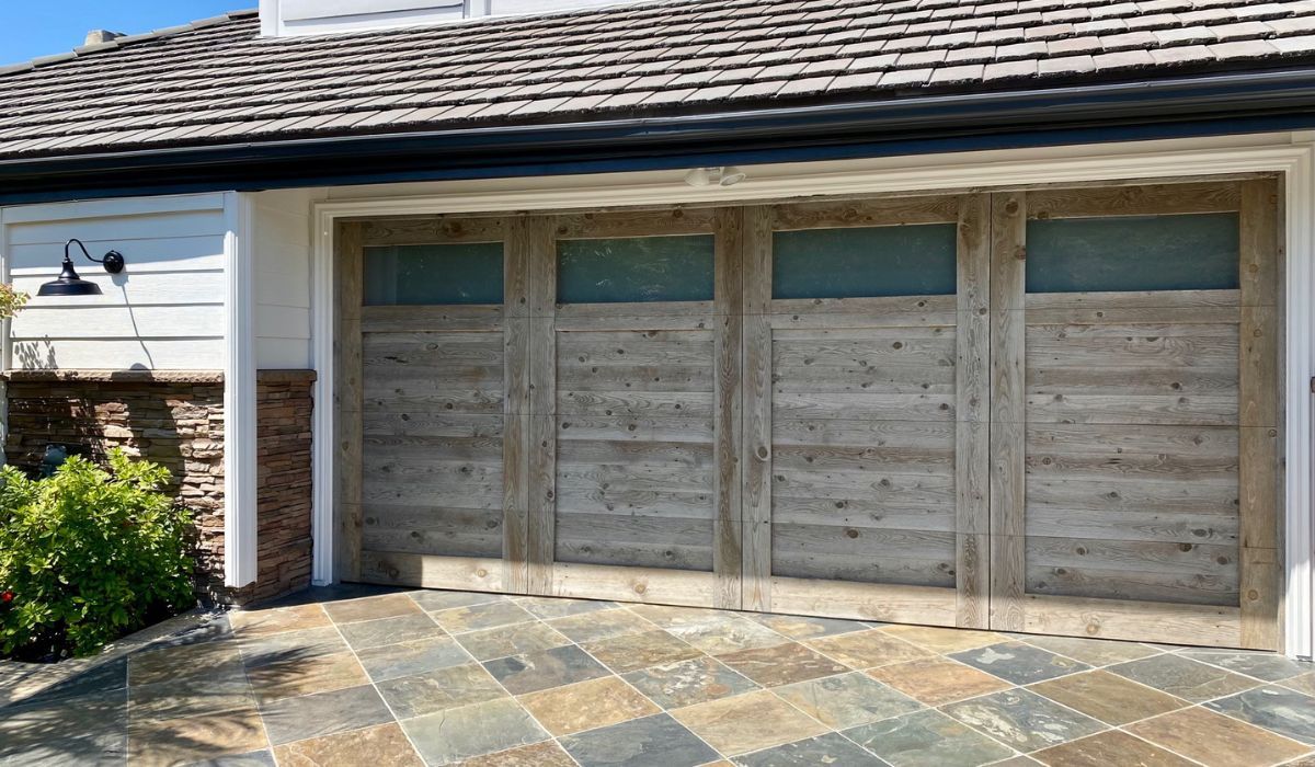 Three-panel weathered wooden garage door with windows on a stone-paved driveway beside a house