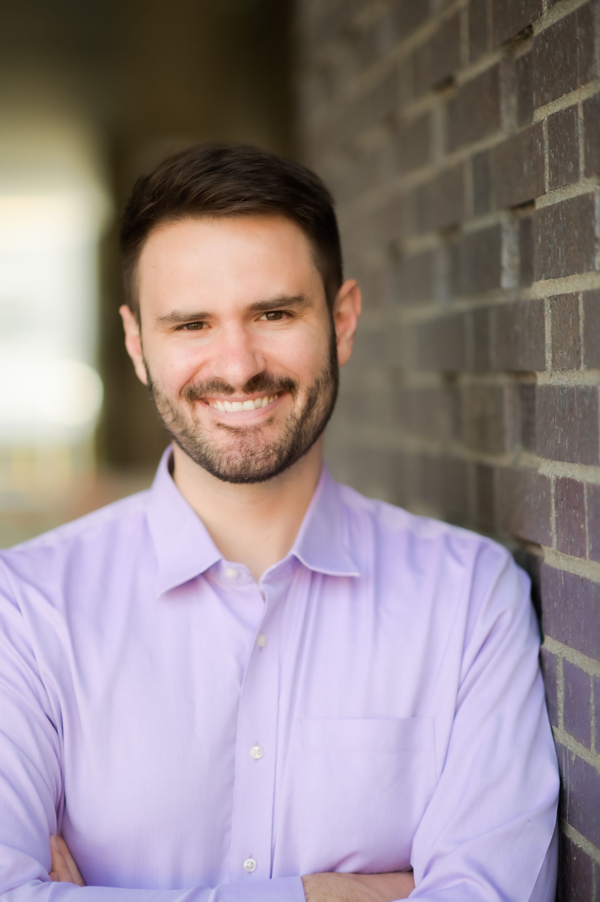 A man in a purple shirt is leaning against a brick wall and smiling.