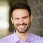 A man with a beard is wearing a blue shirt and smiling.