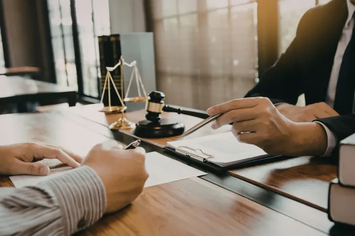 Hands signing a document at a desk during a legal meeting, with a scale and gavel nearby.