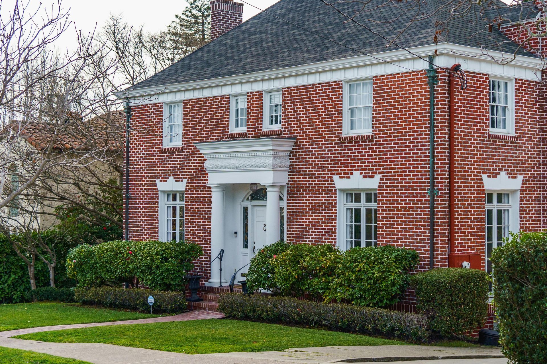 A two-story brick house with a white-pillared entryway and dark shingled roof, surrounded by green hedges.