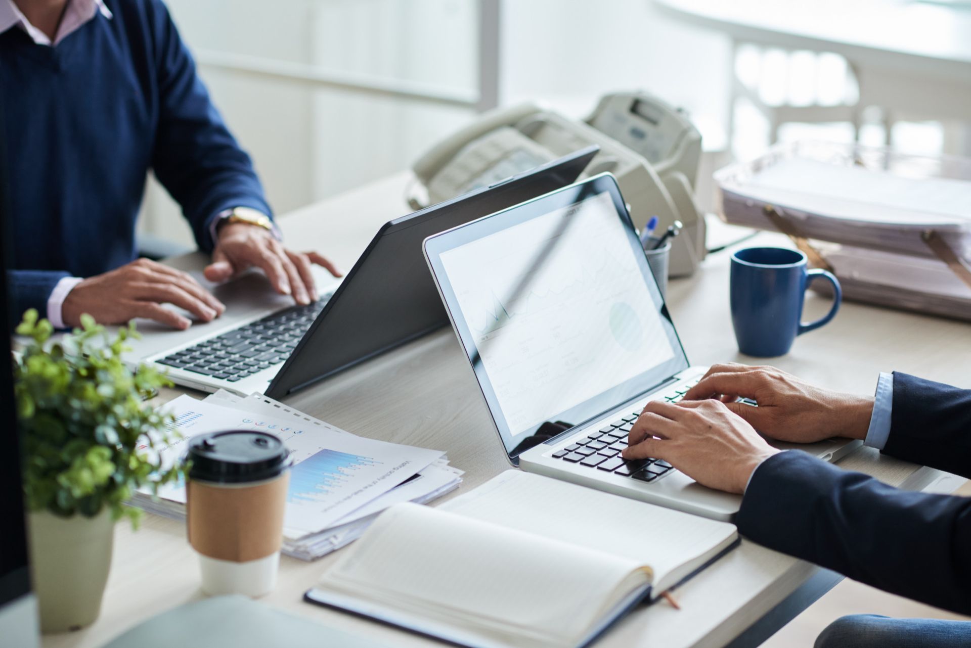 People working on laptops at a shared office table with notebooks and coffee mugs