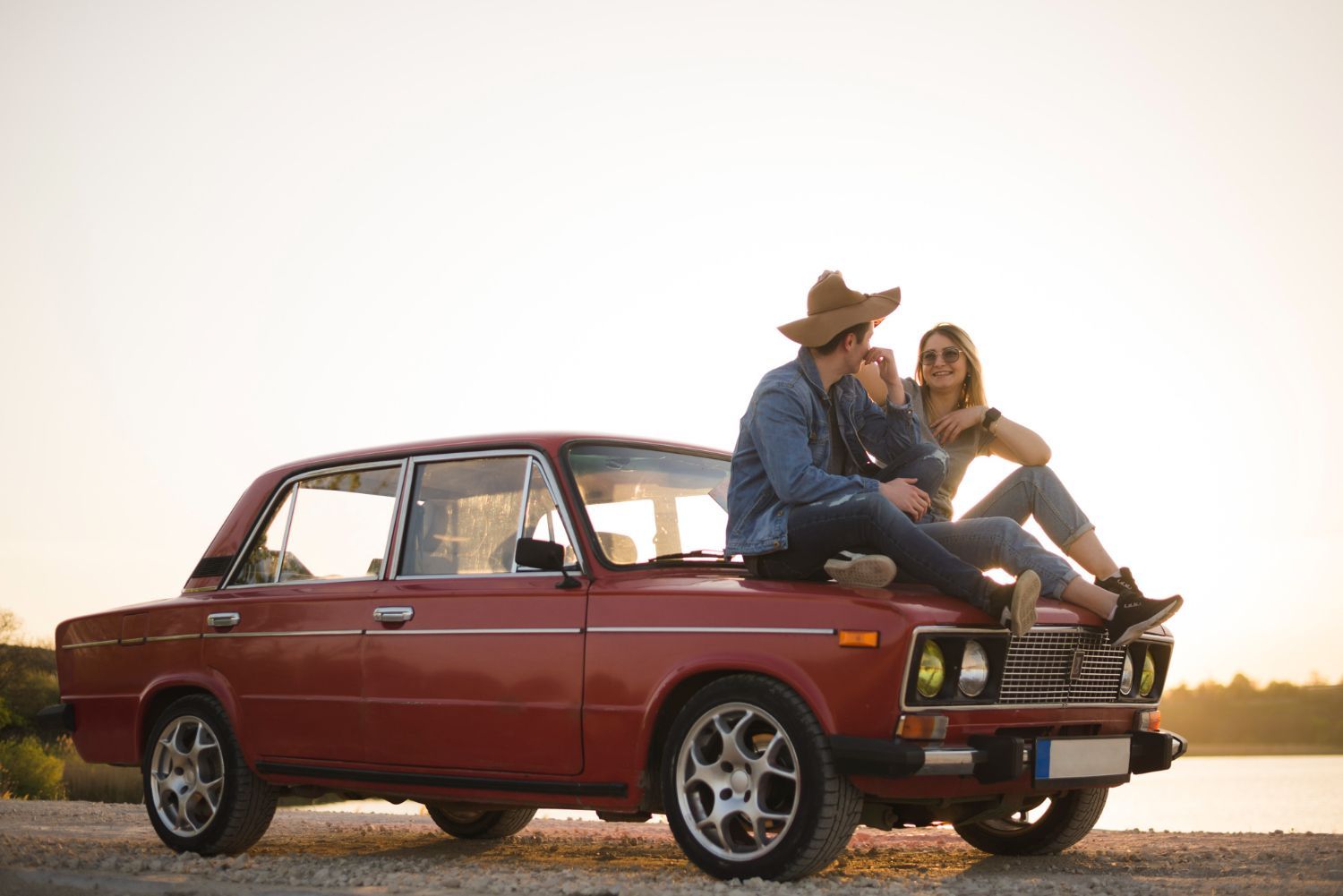 Two people sitting on a red vintage car at sunset by a roadside