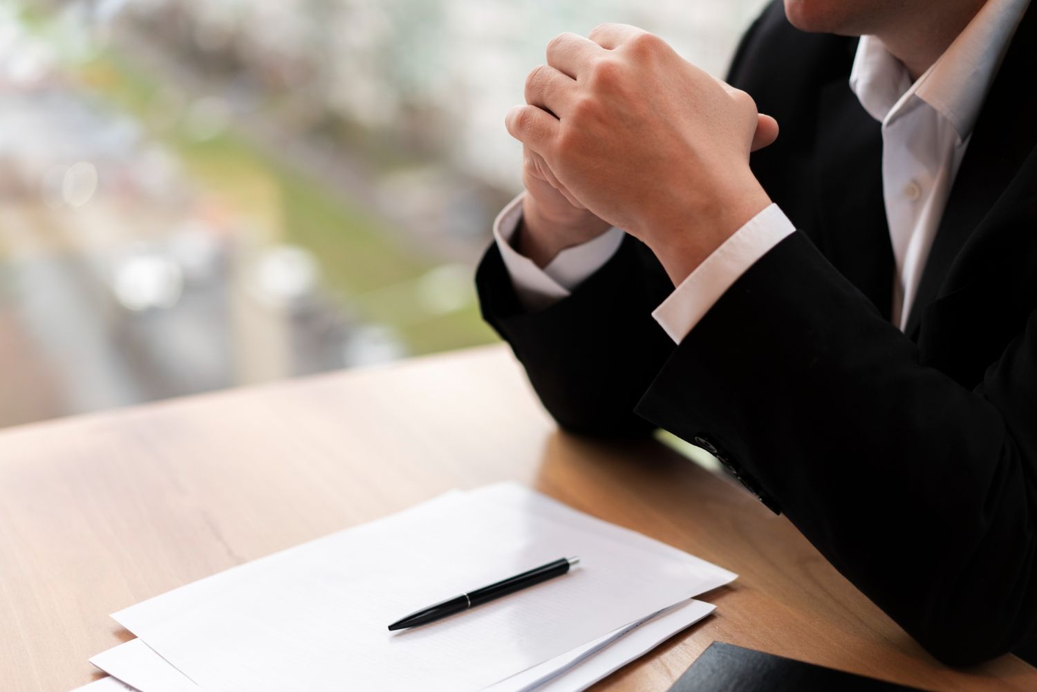 Person in a black suit resting clasped hands on a desk beside a notepad and pen.