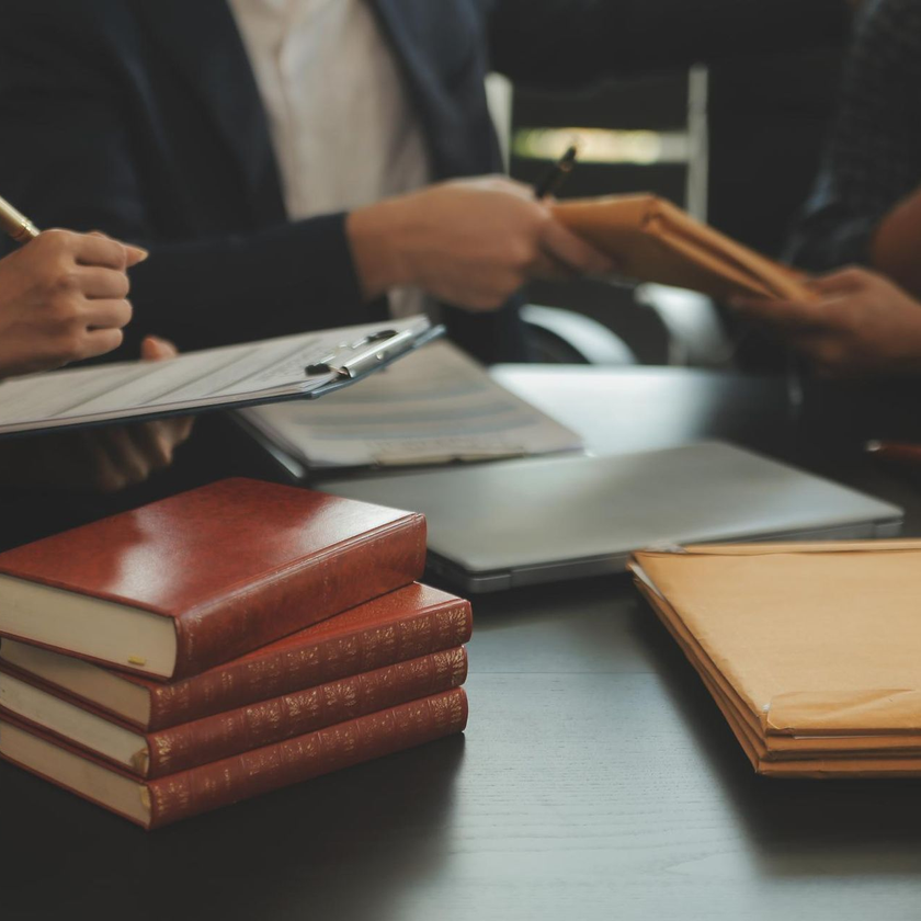 Books, folders, and a laptop on a conference table with people meeting in the background