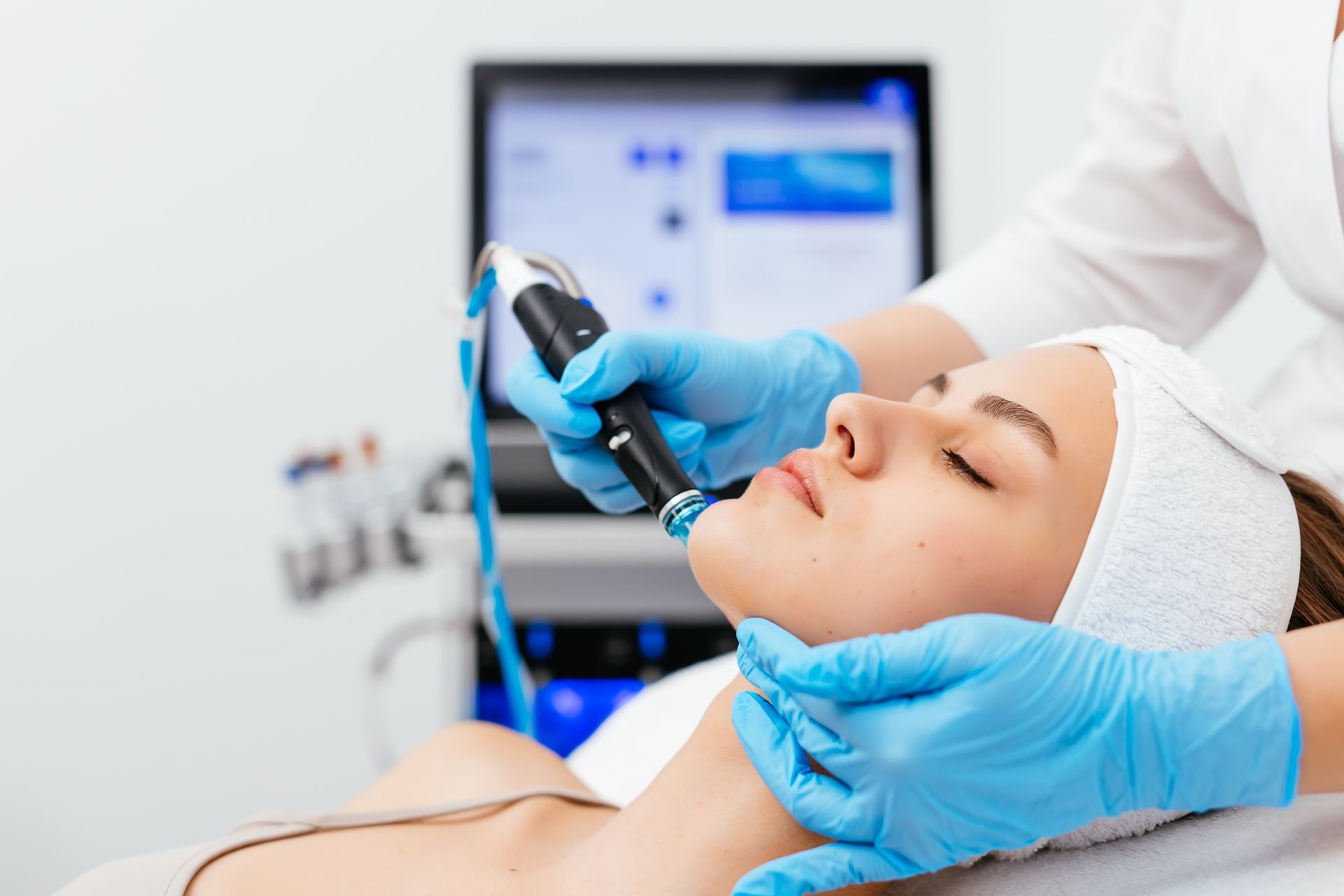 Woman receiving facial treatment at a spa; aesthetician in blue gloves holds a device to woman's face.