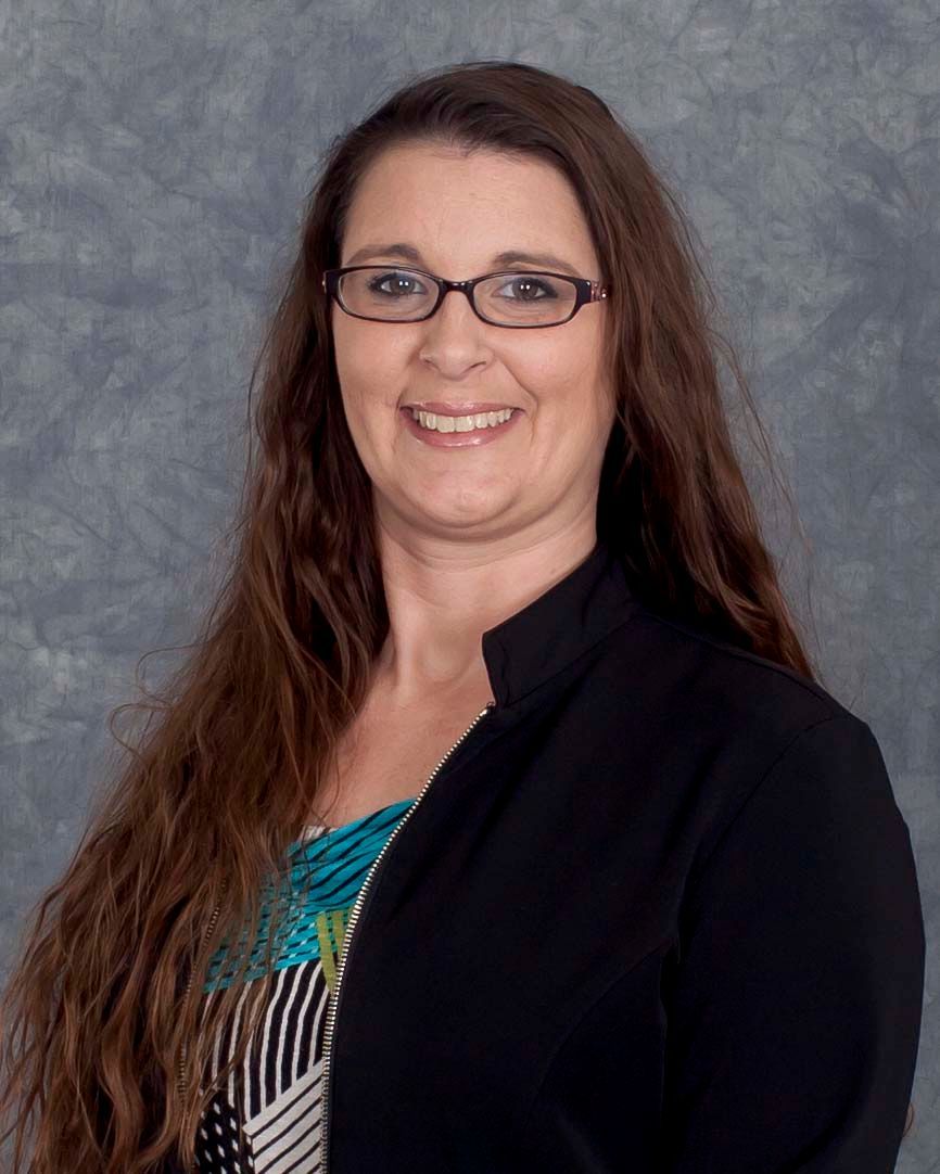 Woman with glasses and long brown hair smiles, wearing a black blazer over a colorful top, against a gray background.