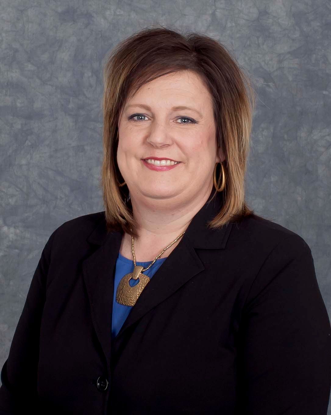 Woman in a black suit smiles, wearing a gold pendant and earrings. Background is grey.