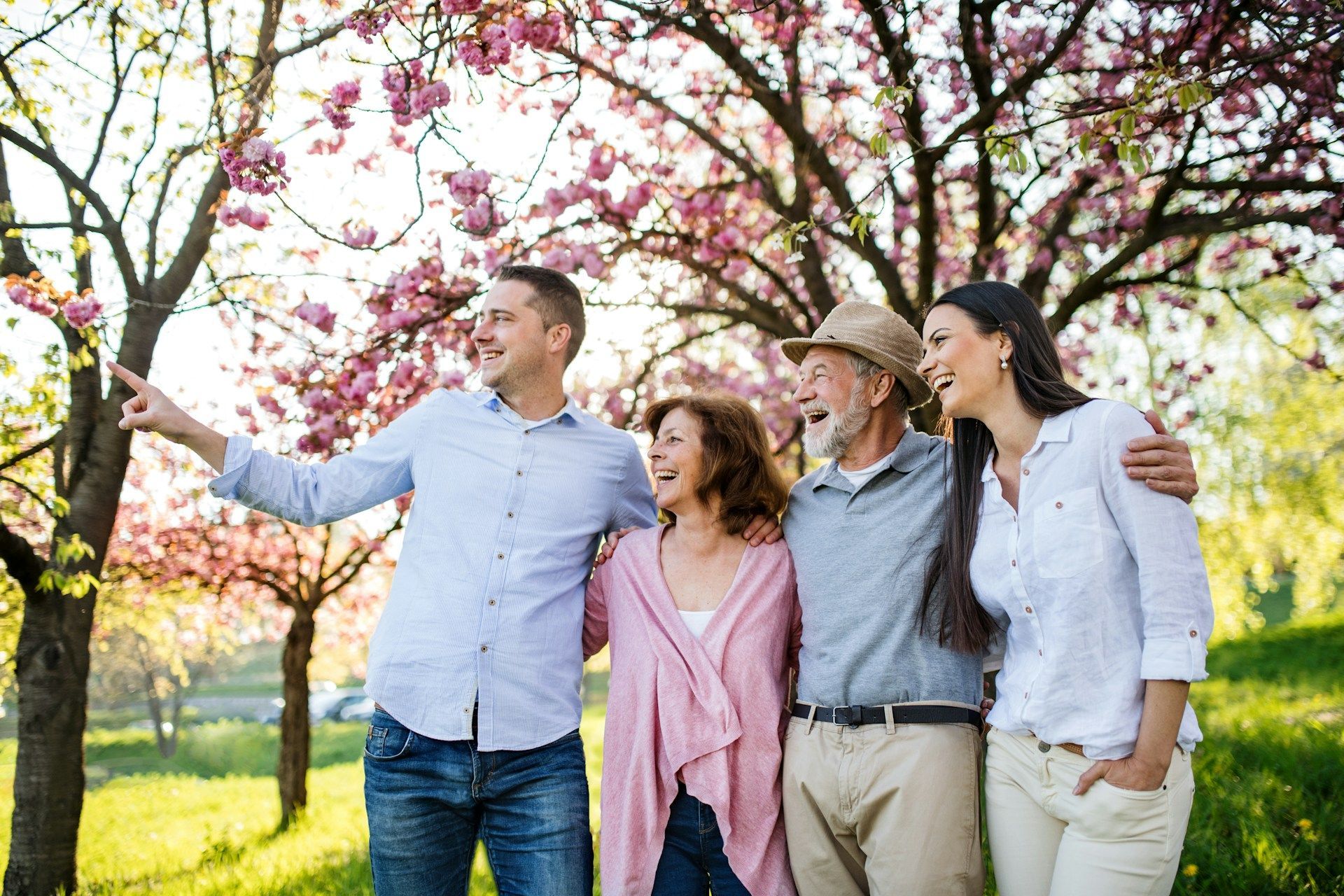 Four people, arm-in-arm, smiling in a park with blossoming trees; one points.