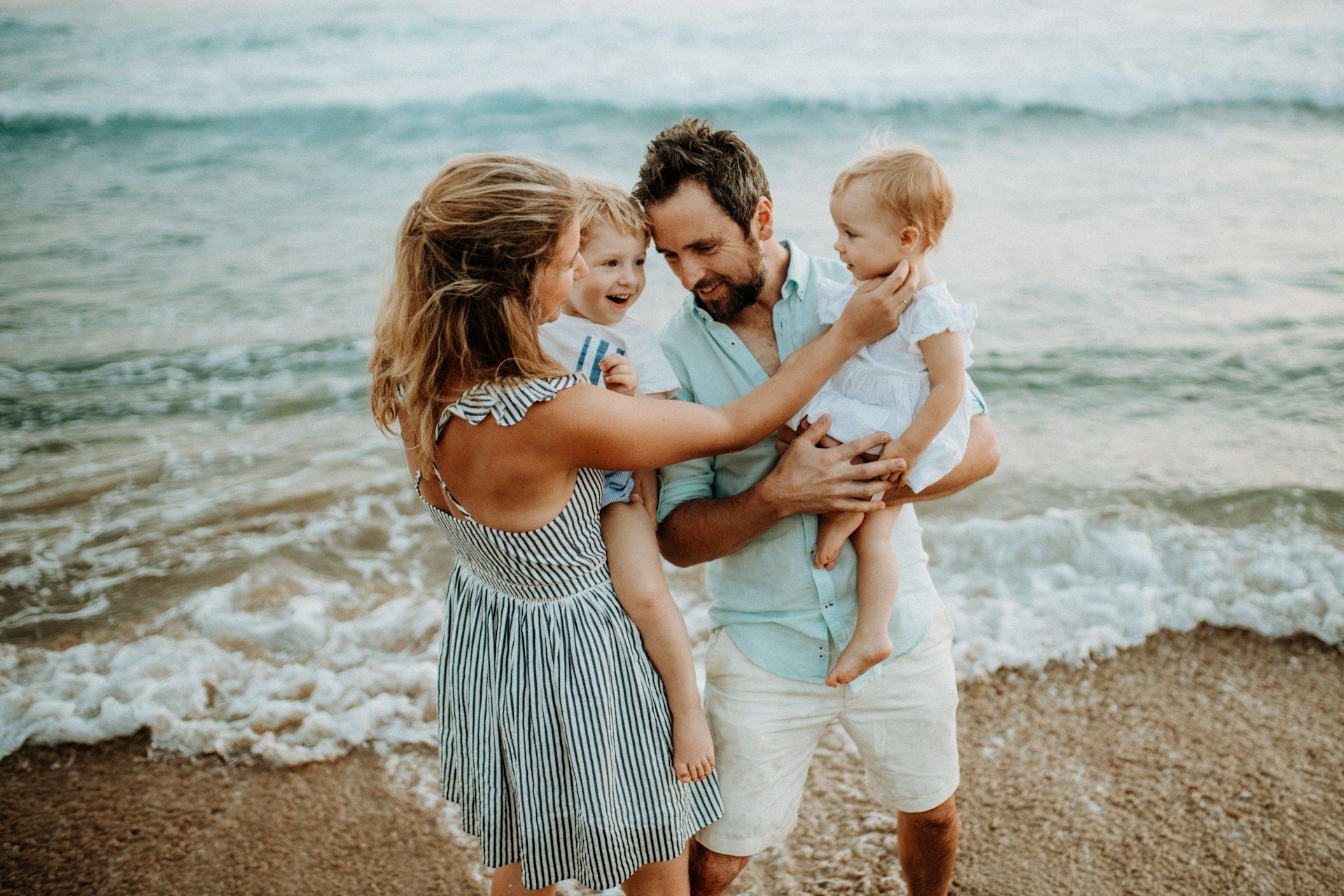 Family of four at the beach, holding two young children. Ocean waves in background.