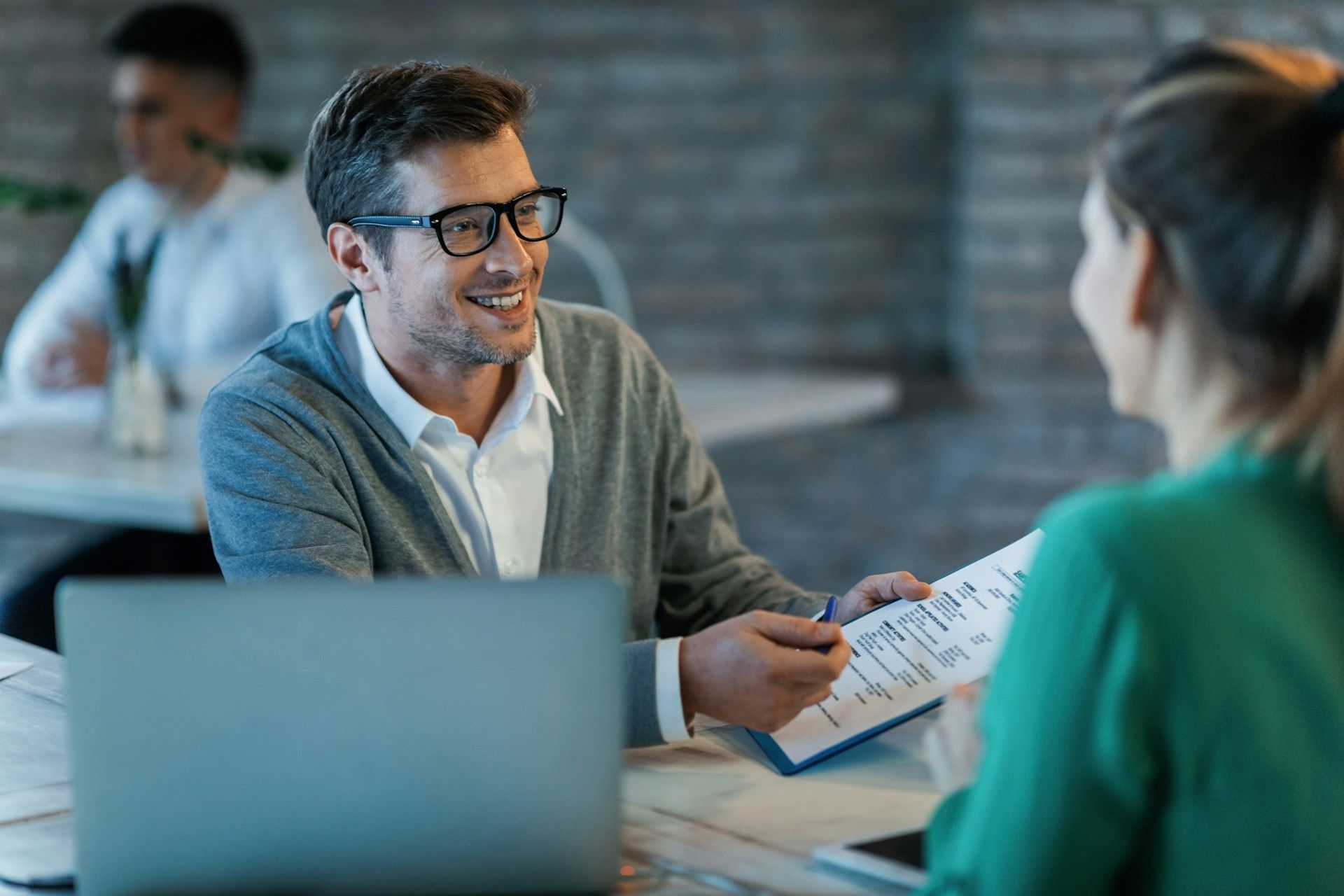 Man in glasses smiling, reviewing documents with a woman at a table, laptop in front.