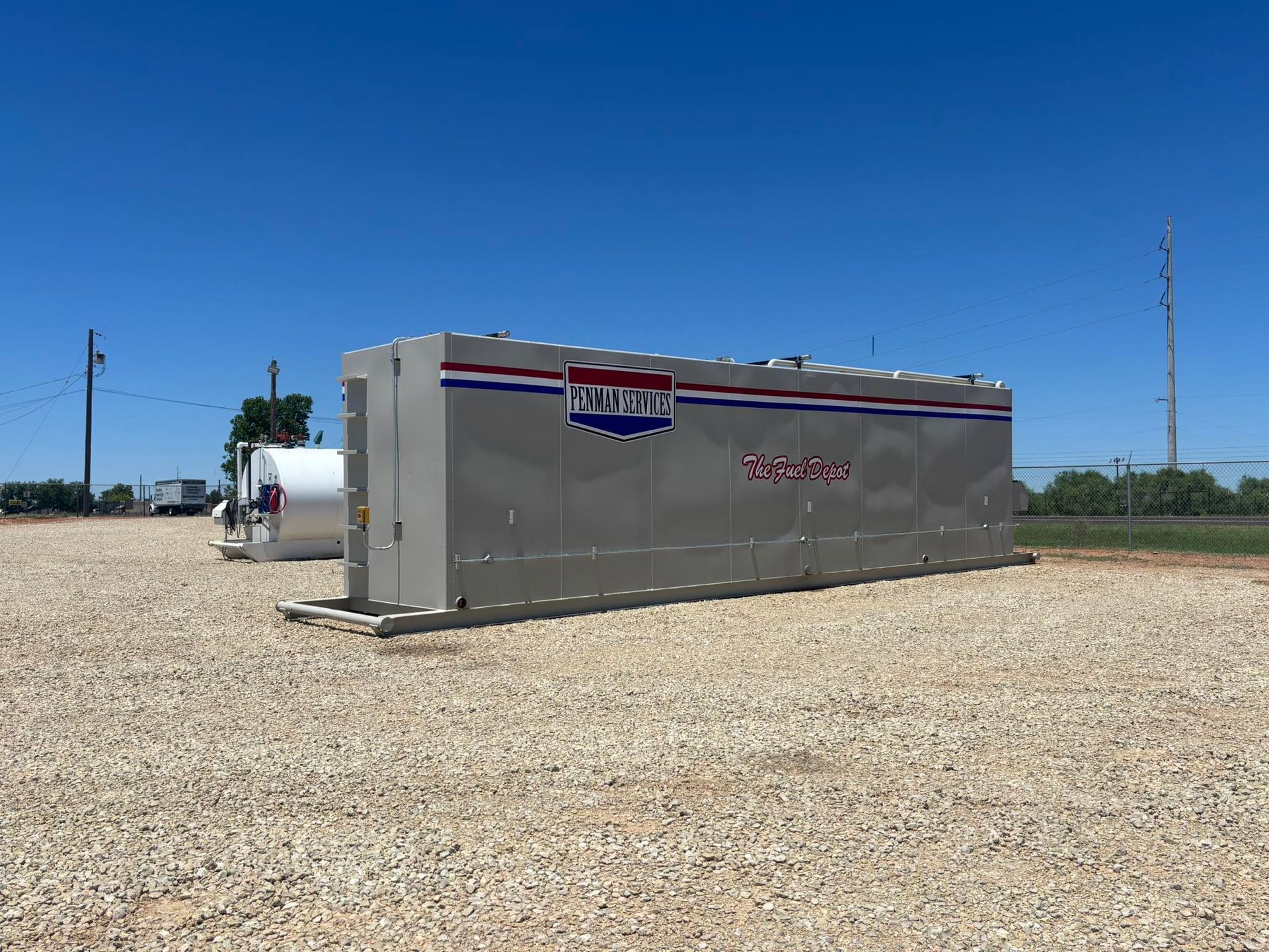 Large silver industrial tank with red, white, and blue logo on gravel under a blue sky.