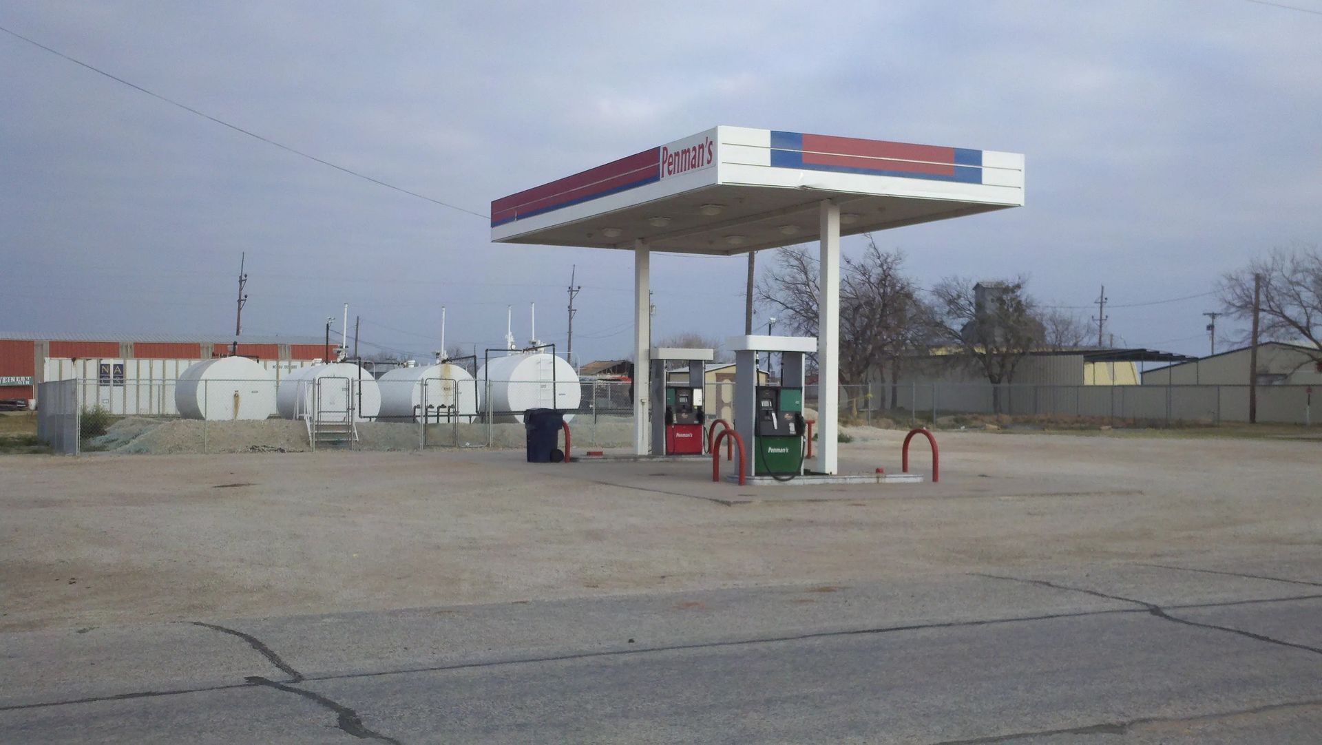 Gas station under a cloudy sky. A canopy covers fuel pumps. Storage tanks and a building are visible.