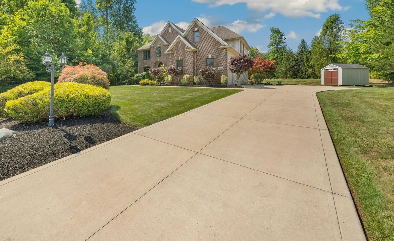 Long concrete driveway leading to a large two-story house with green lawn and trees on a sunny day.