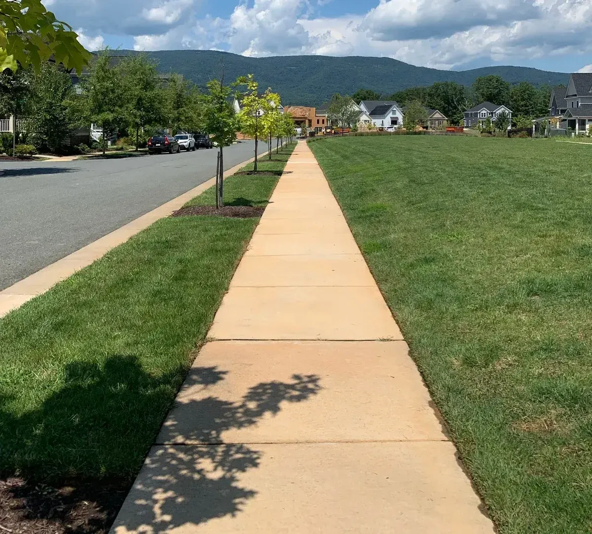 A straight sidewalk through a grassy area with houses and mountains in the background under a sunny sky.