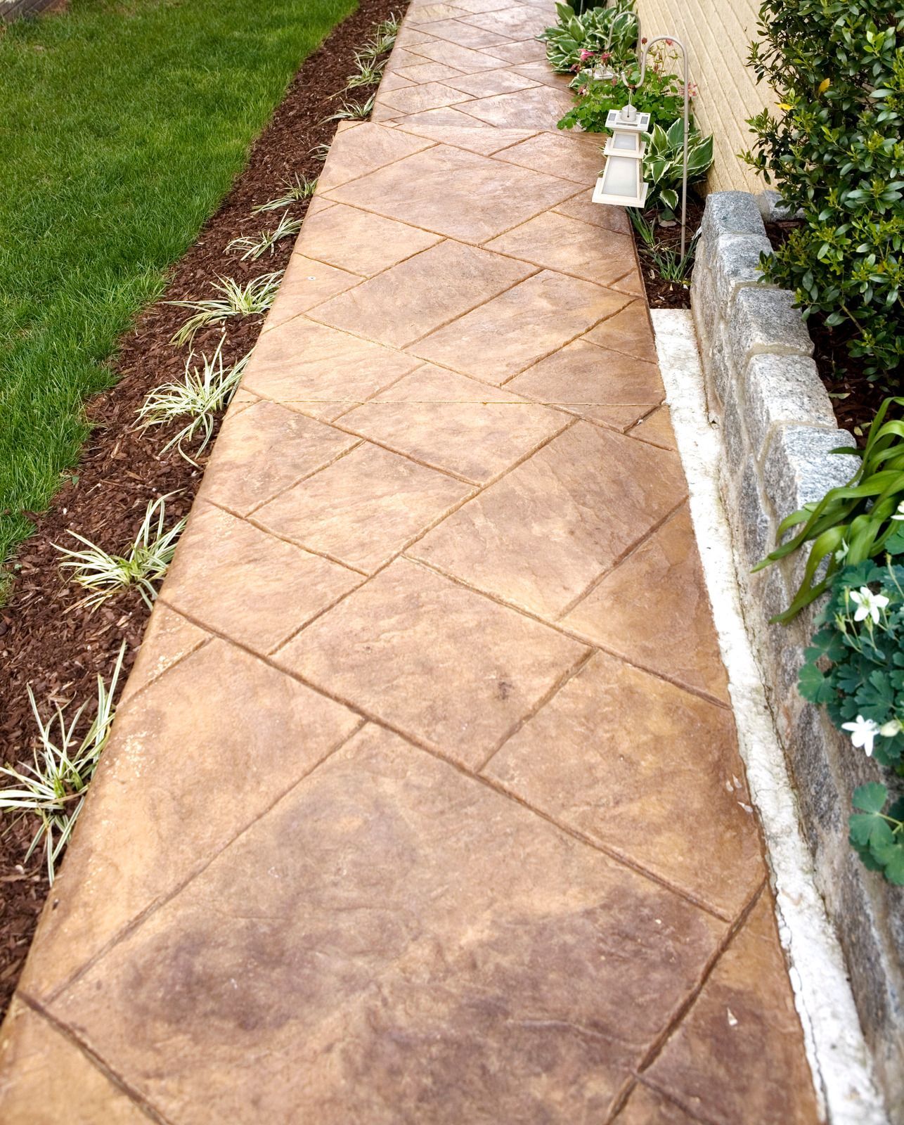 Stamped concrete walkway with diagonal tile pattern, bordered by grass and plants.