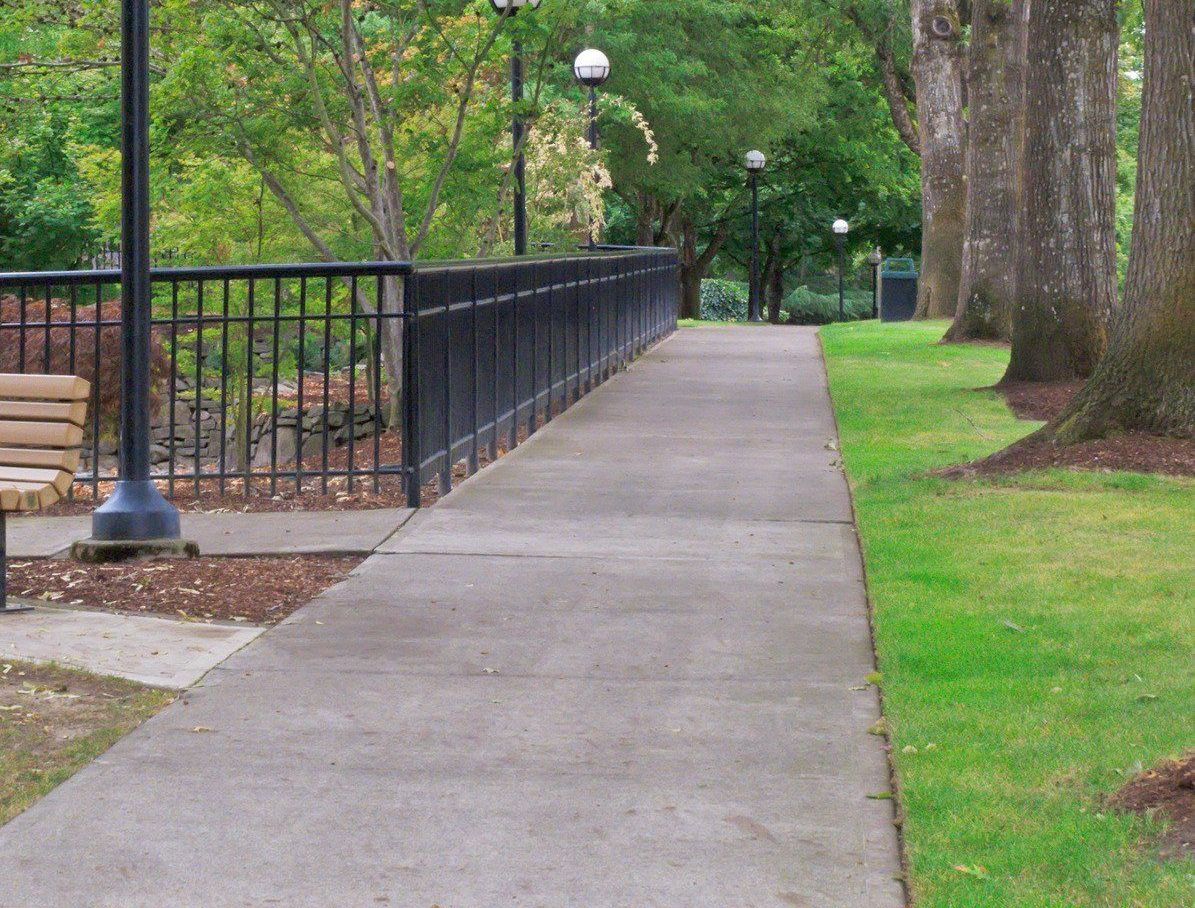 Concrete path in a park lined with black fence, trees, lampposts, and green grass.
