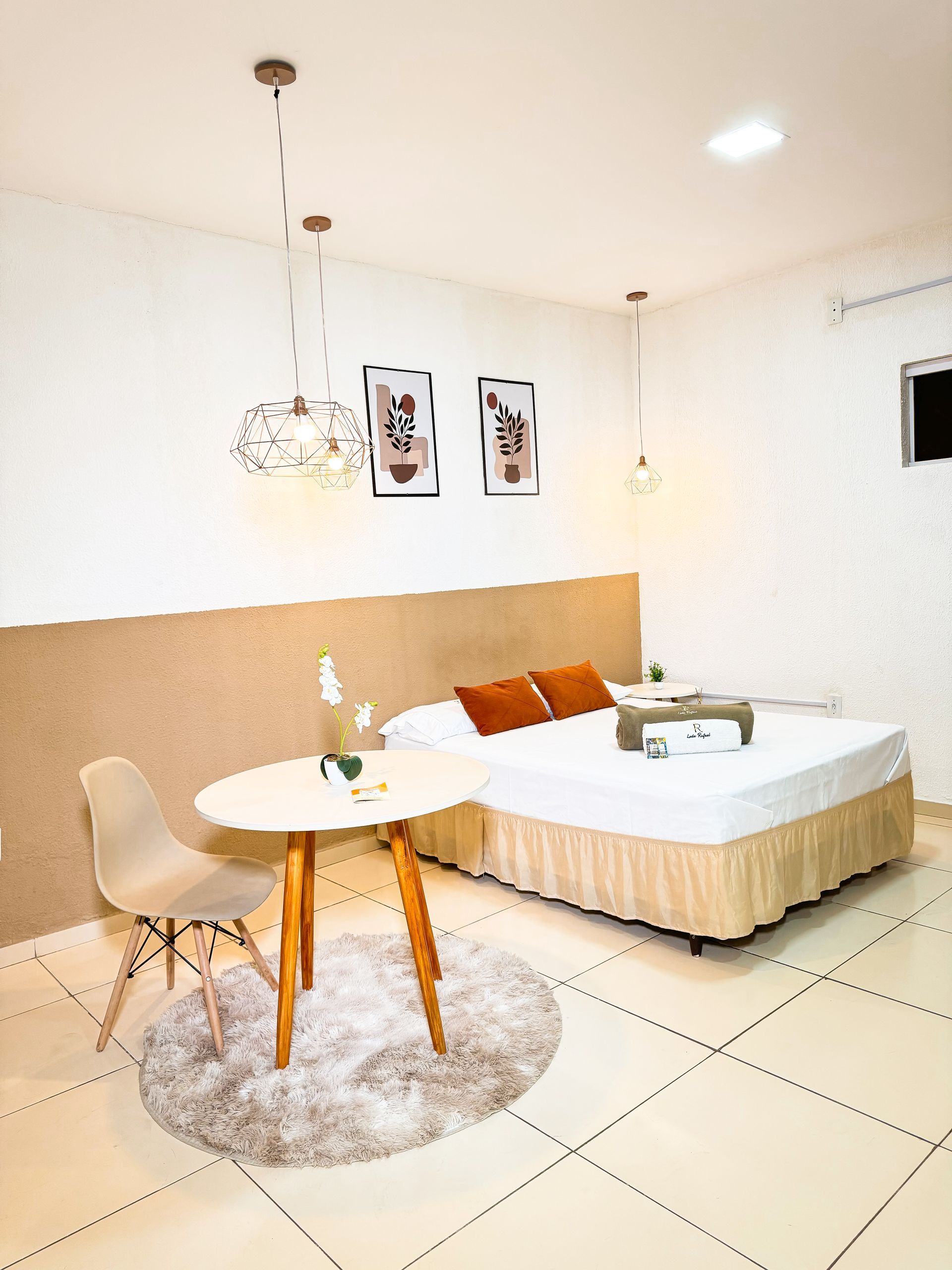 Quarto com cama, mesa, cadeira, iluminação decorativa e duas gravuras emolduradas.