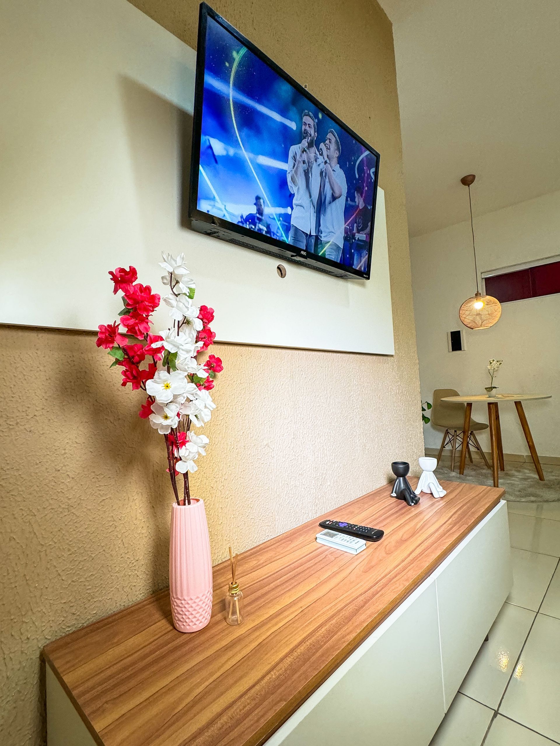 Televisão instalada acima de um armário de madeira com um vaso de flores rosa; mesa de jantar ao fundo.