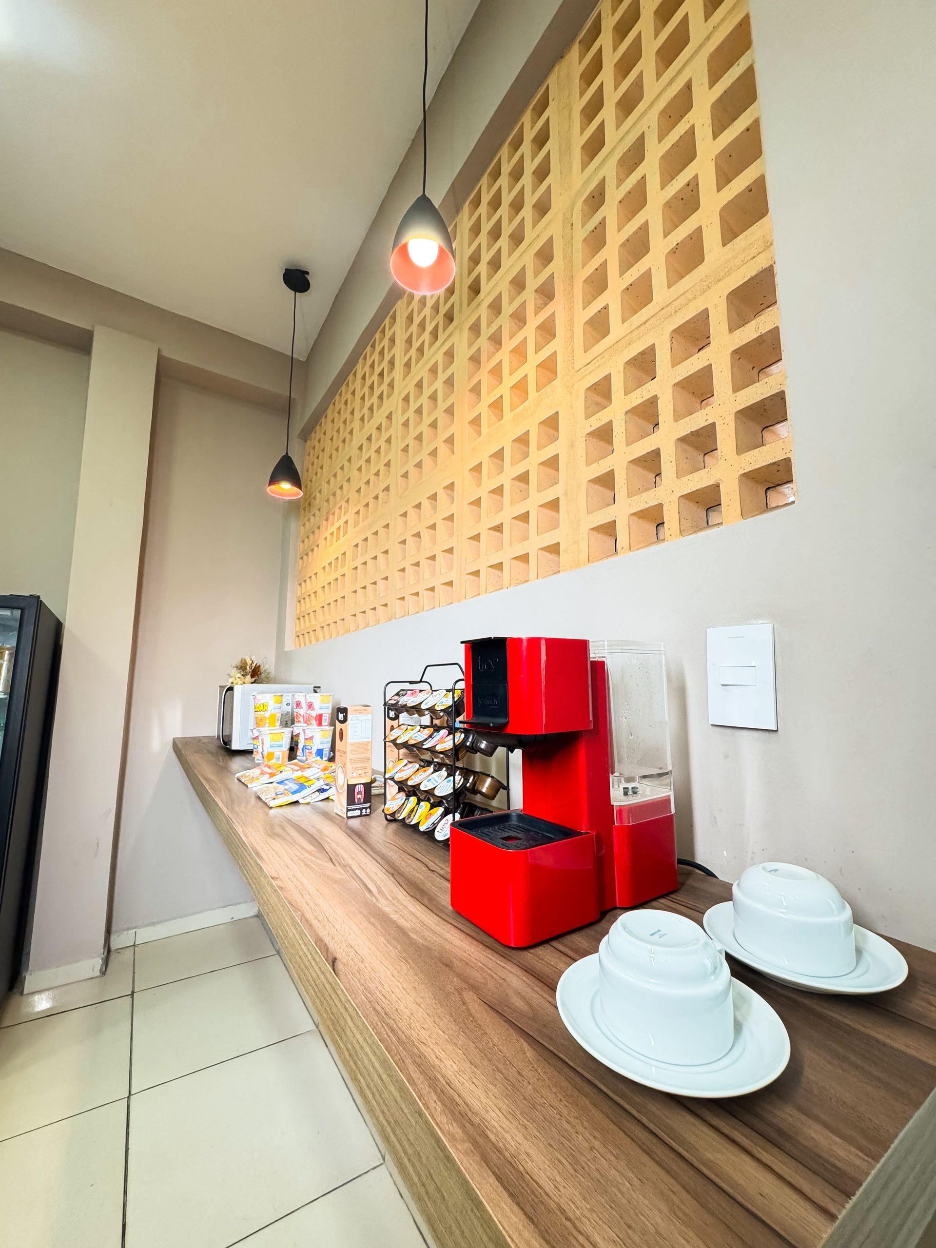 Estação de café com máquina vermelha, xícaras, vinho e parede de tijolos aparentes ao fundo; em um ambiente moderno e luminoso.