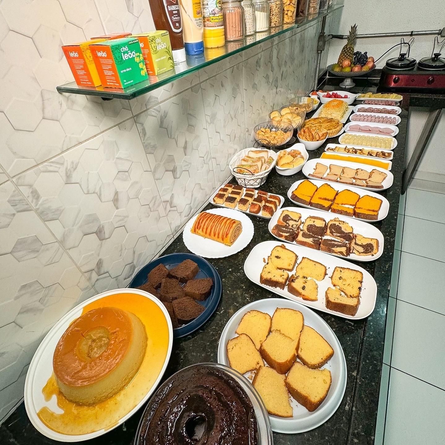 Buffet de café da manhã: Diversos bolos, doces, pães e outros produtos de panificação em um balcão, com condimentos e outros itens em uma prateleira.