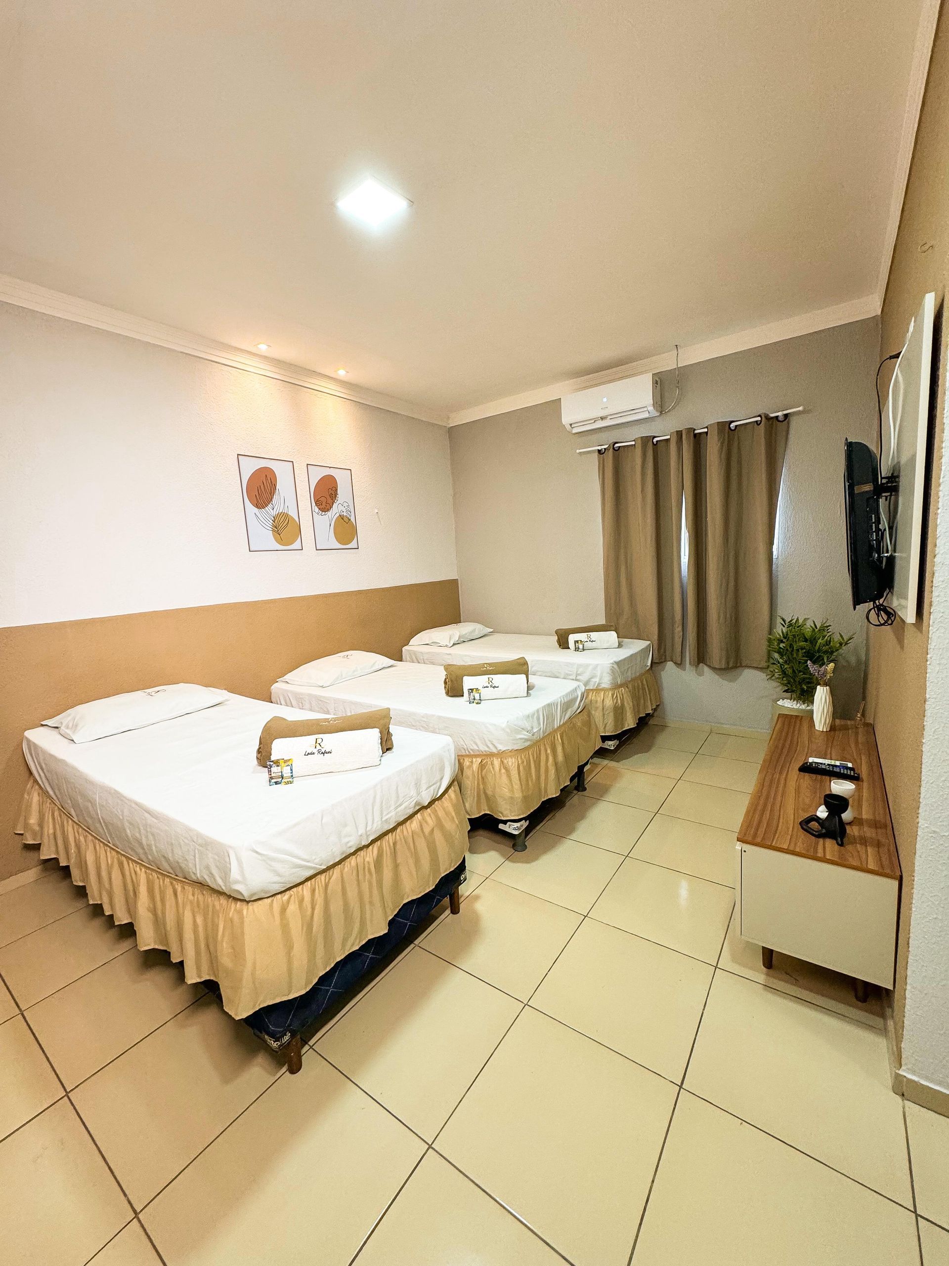 Quarto de hotel com três camas, TV e ar condicionado. Tons de bege e branco predominam.