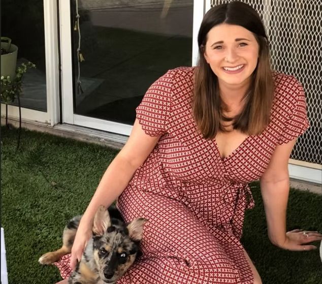 A smiling person with shoulder-length hair wears a reddish-brown patterned dress, sitting on grass beside a spotted dog.
