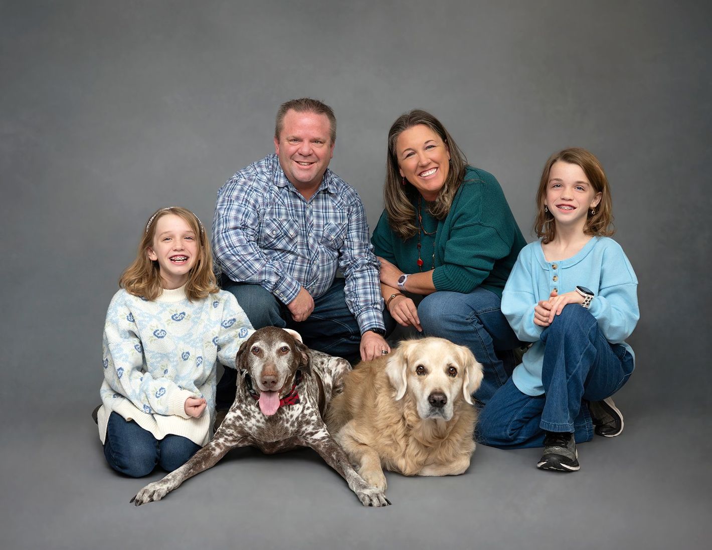 A smiling family of four poses in front of a gray backdrop with their two dogs resting on the floor in front of them.