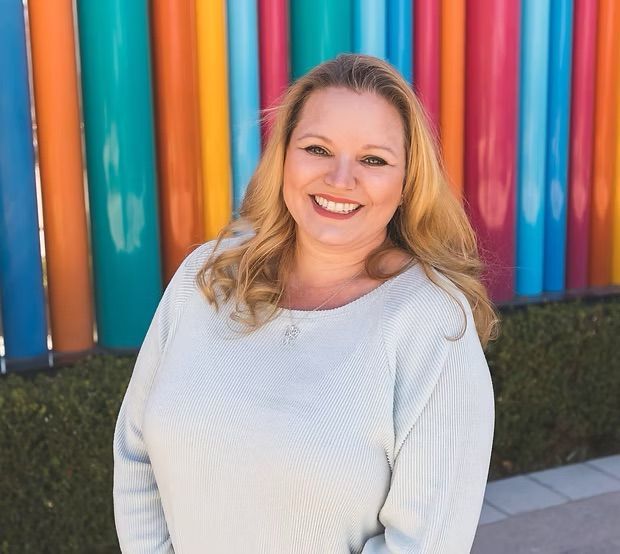 A smiling person with shoulder-length blonde hair, wearing a white sweater, in front of colorful vertical pillars.