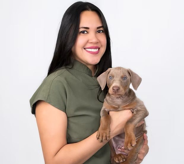 A person with long dark hair, wearing an olive top, smiling while holding a light brown puppy against a white background.