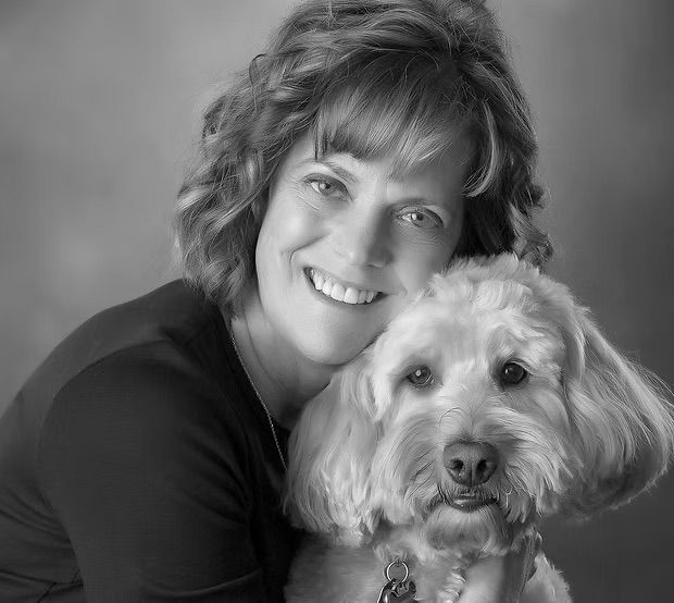 A black-and-white headshot of a smiling woman resting her head against a fluffy, light-colored dog.