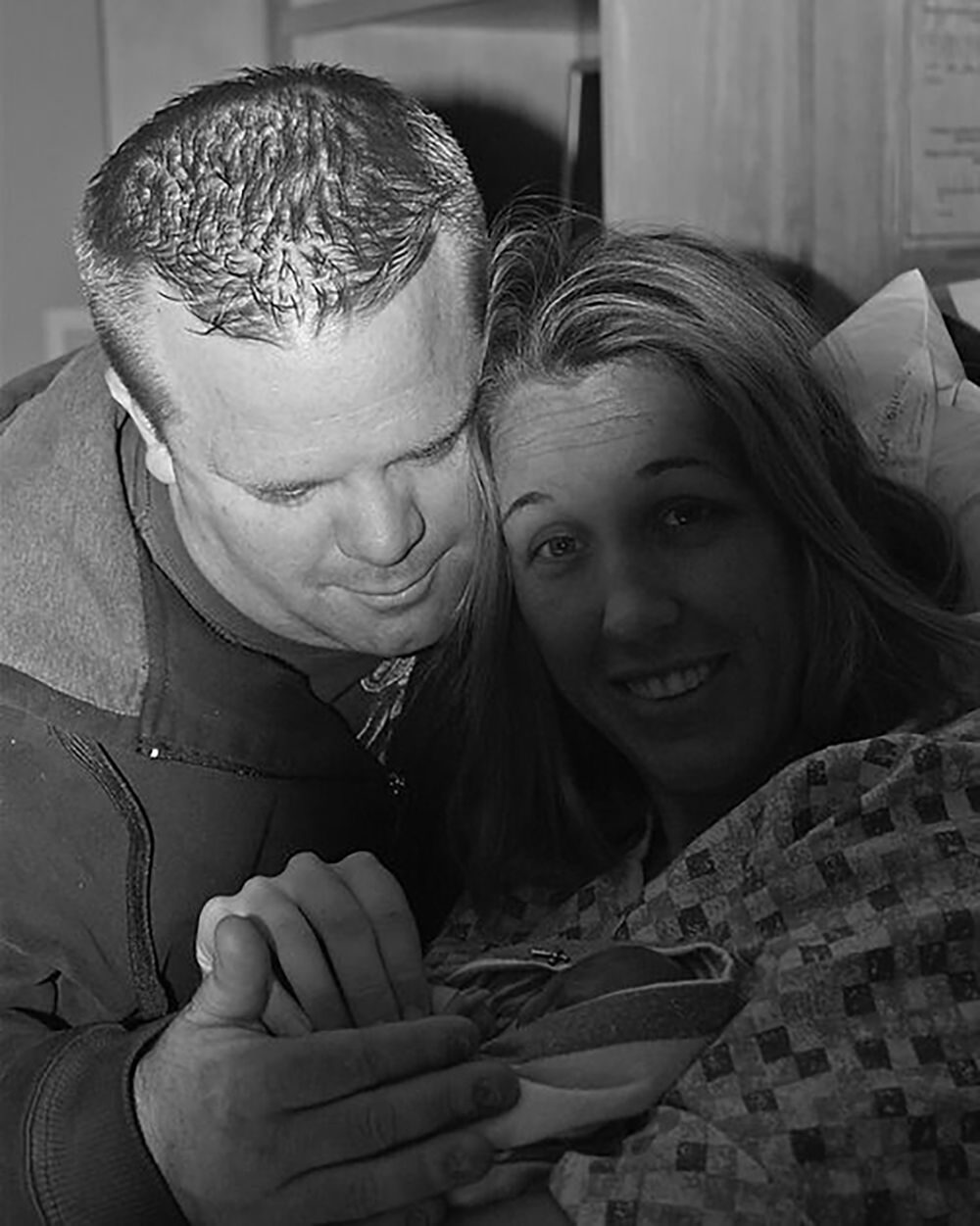 A black-and-white portrait of two people looking down and smiling tenderly at a newborn held in their arms.